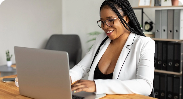 Mulher sentada à mesa em um ambiente de escritório, trabalhando em um notebook. Ela tem pele escura, usa óculos e tem o cabelo trançado solto, caindo sobre os ombros. Veste um blazer branco sobre uma blusa escura e sorri enquanto digita no computador. A mesa é de madeira e, ao fundo, há estantes com pastas e objetos de escritório, indicando um ambiente profissional organizado e bem iluminado.